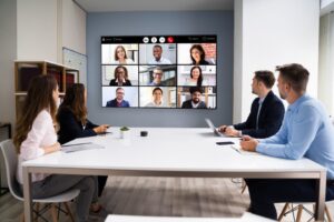 Four individuals gathered in an office or conference room, with a central screen displaying a virtual meeting of nine participants on a conference or Zoom call. This image symbolizes the concept of hybrid work, blending in-person collaboration with remote meetings.