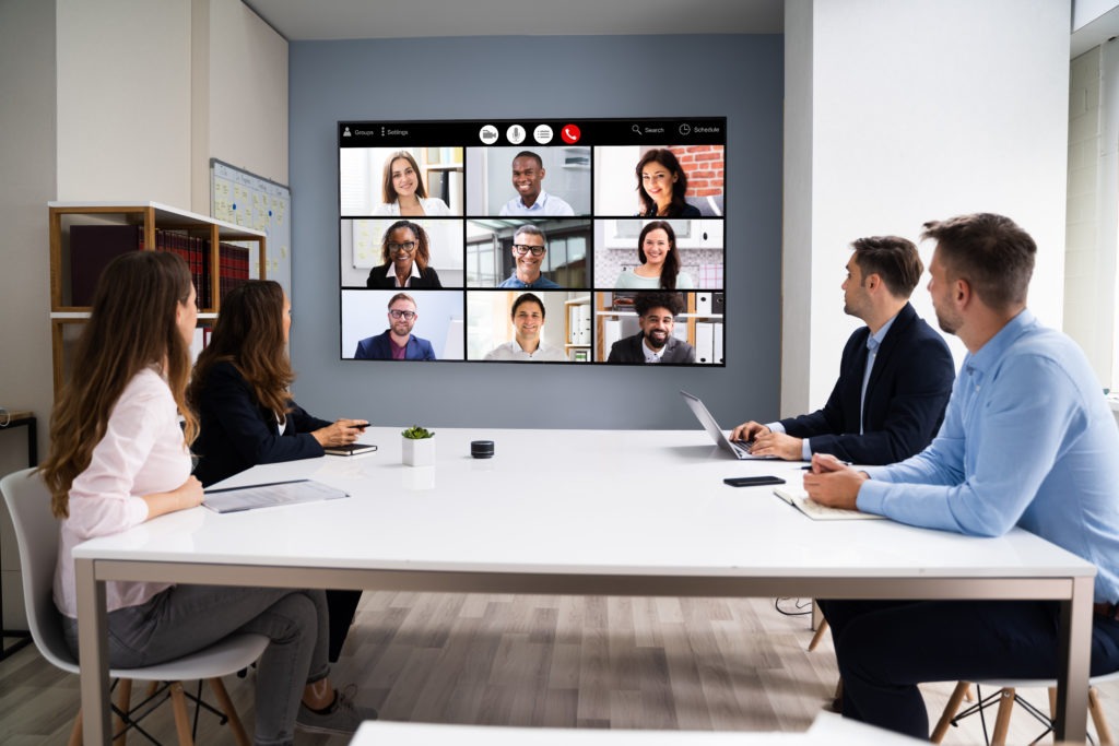 Four individuals gathered in an office or conference room, with a central screen displaying a virtual meeting of nine participants on a conference or Zoom call. This image symbolizes the concept of hybrid work, blending in-person collaboration with remote meetings.